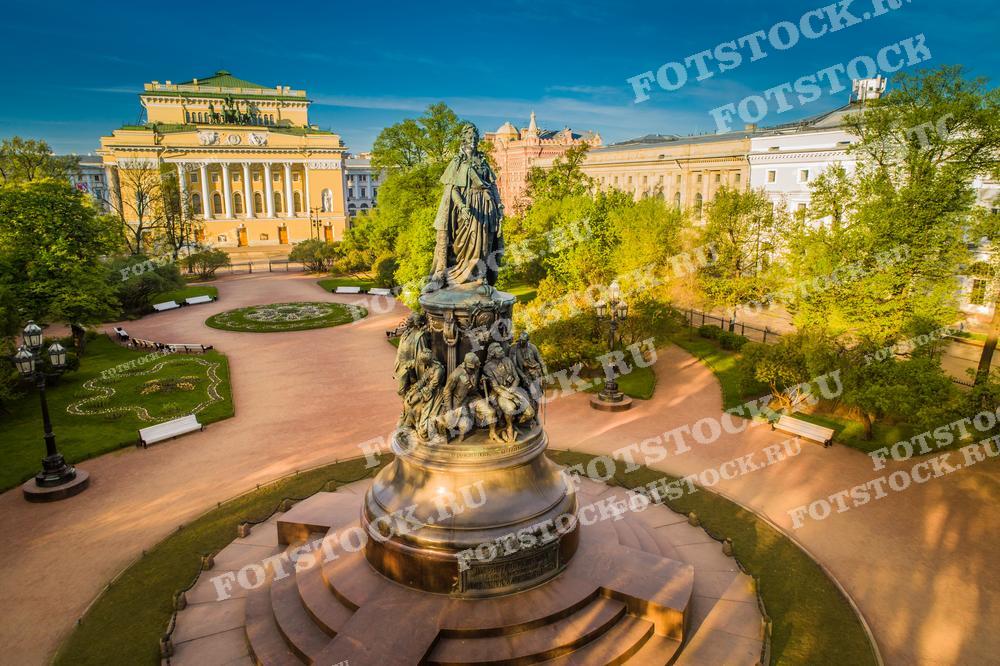 На фотографии изображен памятник Екатерине Великой на фоне Александринского театра в Санкт-Петербурге. Этот вид показывает пышную зелень деревьев и аккуратные клумбы парка Островского, где расположен монумент. Мягкий свет утреннего или вечернего солнца придает сцене особое очарование. Это изображение подойдет для исторических материалов, туристических гидов, культурных обзоров или в качестве декоративного элемента в различных публикациях о России и её культурном наследии.